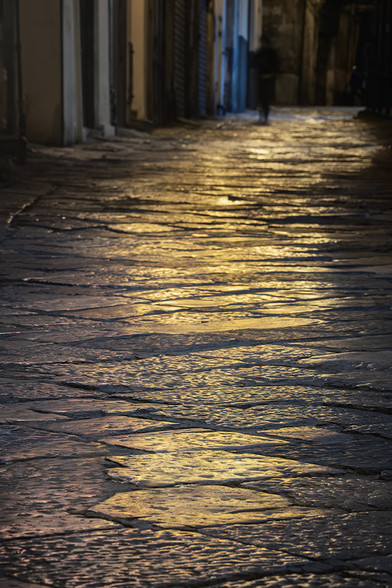 Streetlights create a golden glow at night on the ancient cobblestones in a narrow alleyway in Palermo Sicily. A solitary figure is seen in the distance, adding a sense of solitude to the scene.