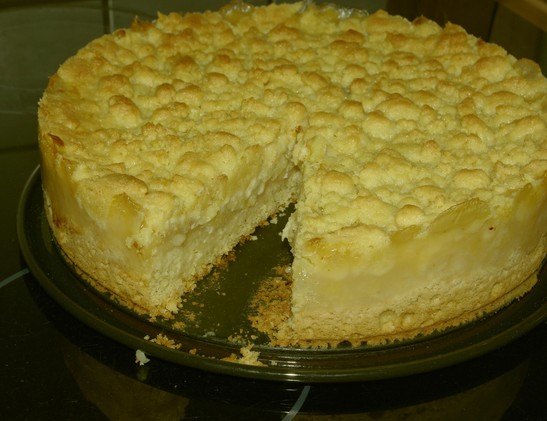 frischer Streuselkuchen. Der Boden sieht weich und fluffig aus, darauf eine Apfelmassefüllung, darüber eine Decke aus Kuchenstreuseln. Der Kuchen sitzt auf dem Unterteil einer Back-Springform. Ein Kuchenstück fehlt schon. Dadurch kann man das Innere des Kuchens sehen.