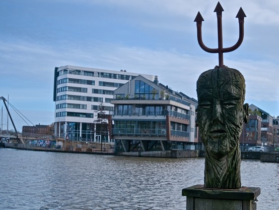 View of a harbour. The face of a man made of weathered wood can be seen in the foreground. A trident protrudes from his head. It is a sculpture by artist Georg Willms. Behind him you can see various houses, some of which protrude into the water. Many windows offer a great view of the water. The sky is slightly cloudy, but there is also a lot of blue to be seen. The colours are cold, it's a January day.