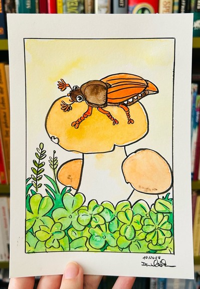 Foto: Meine Hand hält ein Aquarellbild hoch, darauf ist eine Comicillustration von einem Maikäfer, der auf einen Pilz sitzt. Unter dem Pilz sind ganz viele Kleeblätter.