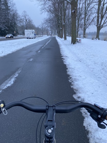 Blick über einen Fahrradlenker auf einen breiten, asphaltierten Radweg. Die Landschaft ist verschneit, links einige Autos auf einer Straße. Der Radweg ist geräumt.