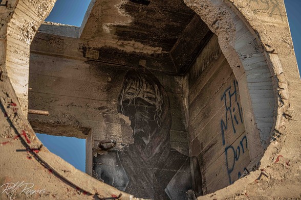 A graffiti painting of a woman's face, masked, on the interior wall of an abandoned building, peering thru the circular opening to the outside.