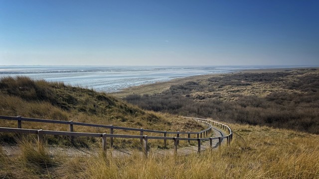 A wide-angle view from a vantage point overlooking the Wadden Sea and the dune landscape of the island of Ameland. A winding path, secured with a wooden railing, leads from the viewpoint to the Wadden Sea. The weather is sunny, bright and friendly with a blue sky.