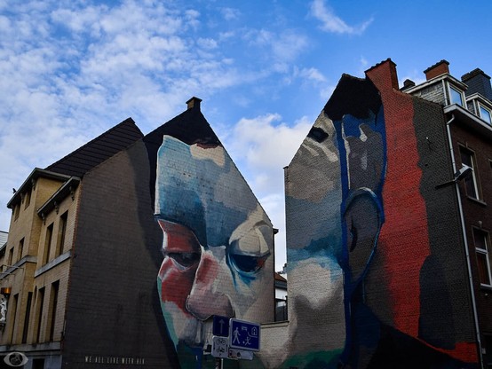 Place of a tornn down corner house revealing the two facades of the attached houses. On both facades is a street art picture painted in red, blue and white blotches that together make the picture of a face. On the left facade at the bottom is written the words ‘we all live with HIV.'