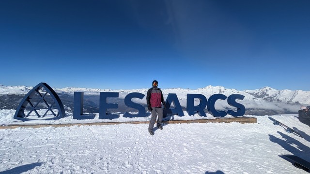 I stand in the snow in front of the Les Arcs sign at the top of the mountain. The sky is blue and there are more mountains in the background