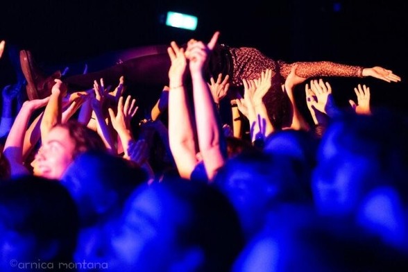 A person crowd surfing at a concert, supported by many hands, with a vibrant blue and purple lighting illuminating the scene. The crowd appears energetic and engaged.