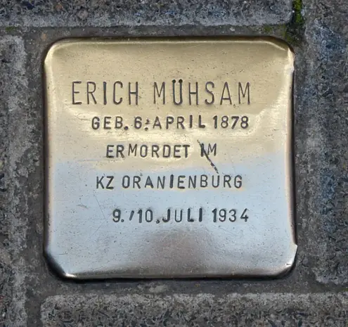 The image shows a brass memorial plaque, known as a "Stolperstein" or "stumbling stone," dedicated to Erich Mühsam. The plaque includes the following information:

- His name: Erich Mühsam
- Date of birth: April 6, 1878
- The fact that he was murdered in the concentration camp at Oranienburg
- The date of his death: July 9/10, 1934

These stones are placed in

Image Credits: Wikimedia / Ralf Küpper / CC BY-SA 3.0 de