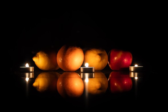 A series of fruits (two lemons, an orange and an apple) are seen under the light of candles. They are reflected in the surface they rest on.