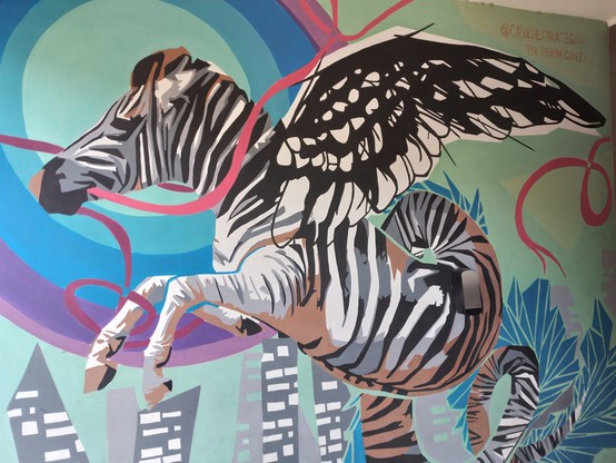 A colorful blue and pink graffiti in a wall of a building. It depicts some kind of zebra with wings and serpent body.