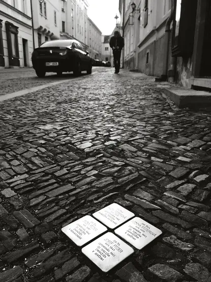 The image appears to show a cobblestone street with a person walking in the background. On the ground, there are several small white stones or plaques, likely memorials, inscribed with text. The overall tone of the image is black and white, contributing to a somber atmosphere.

Image Credits: Wikimedia / Francisco Peralta Torrejón / CC BY-SA 4.0