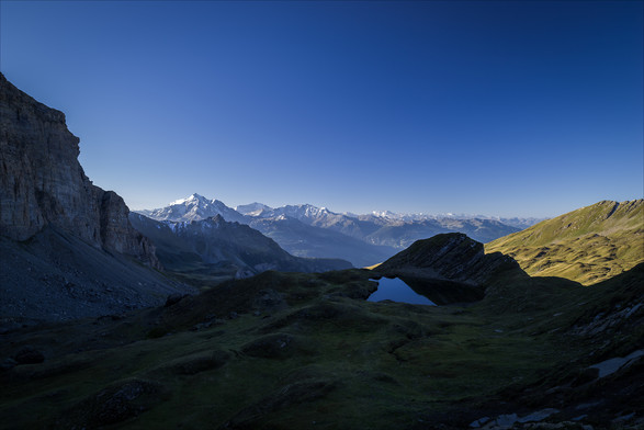 Photo d'un paysage de montagne.
Une petite vallée encaissée est plongée dans l'ombre. Le côté gauche de l'image est fermé par une haute falaise qui domine un pierrier. Plus à droite on arrive sur un pâturage tourmenté de reliefs arrondis laissés par le retrait d'un glacier peut-être récent. Au bout de ce pâturage se trouve un petit lac niché au creux d'une formation rocheuse en forme d'écaille. A droite derrière cette écaille il semble y avoir une rupture de pente brutale, on voit au loin un pâturage aussi tourmenté, et éclairé par le soleil. A l'horizon on distingue de hautes montagnes aux sommets enneigés, de l'autre côté de la vallée. Au-dessus, le ciel est d'un bleu immaculé.
-
Photo of a mountain landscape.
A small, deep valley is plunged into shadow. The left side of the picture is closed by a high cliff overlooking a scree slope. Further to the right, we come to a pasture tormented by rounded reliefs left by the retreat of a perhaps recent glacier. At the end of this pasture is a small lake nestling in the hollow of a scale-like rock formation. To the right, behind this scale, there seems to be a sharp break in the slope, and in the distance we can see a similarly tormented, sunlit pasture. On the horizon, high, snow-capped mountains can be seen on the other side of the valley. Above, the sky is immaculately blue.