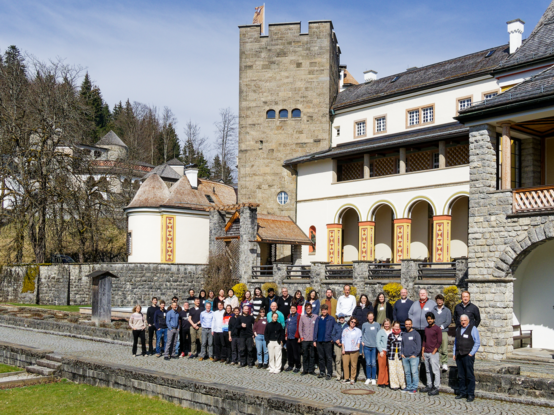 Scientists involved in the Q-Arctic project posing for a group photo in front of Schloss Ringberg, a historic stone building with arched windows and a tower surrounded by trees and a cobblestone pathway under a clear blue sky.