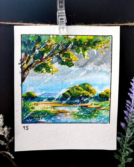 Square watercolor framed with a black line on a rectangular, portrait-oriented paper. A stormy spring scene unfolds as raindrops streak through the air, refreshing the lush greenery. Dark clouds loom above, yet patches of blue sky peek through, casting a shifting light over the rippling water. A large tree branch in the foreground frames the view, its leaves dripping with rain. Distant mountains fade into the mist, adding depth to the moody, rain-soaked landscape. Number 95 is written in the bottom left corner.