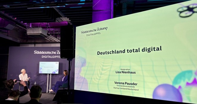 Ein großer Screen zeigt den Titel der Veranstaltung im Hintergrund des Raumes sind zwei Speakerinnen zu sehen.