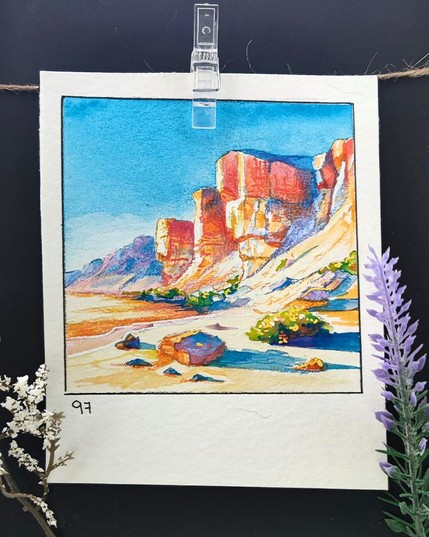 Square watercolor framed with a black line on a rectangular, portrait-oriented paper. A desert landscape with towering red and beige rock formations under a bright blue sky. The foreground features scattered stones, a sandy path, and patches of green vegetation with small flowers. In the bottom left corner, the number 97 is written.