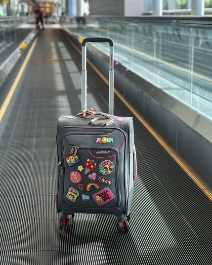 A wheeled suitcase with various colorful stickers and patches, displaying the word "ALOHA" and images like flowers and rainbows, is placed on an airport moving walkway. The modern, spacious terminal is visible in the background.