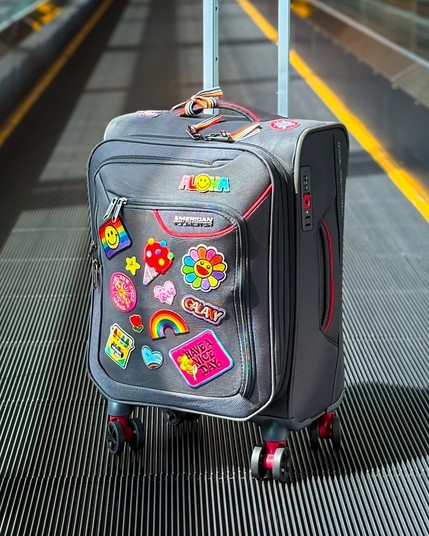 A grey rolling suitcase adorned with colorful patches, including smiley faces, flowers, ice cream, and a rainbow. It has a sturdy handle and wheels, set against a travelator in an airport area.