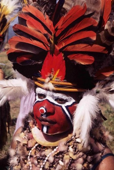 An indigenous person with face painted red & white around the eyes with a headdress made from red bird of paradise feathers and white plumes on the ears.