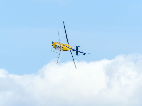 A yellow and blue helicopter, positioned vertically instead of horizontally in the air, as it banks in to a sharp turn. The helicopter is frame by blue sky and fluffy white clouds