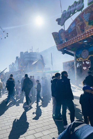 A portrait-format scene on a big fun fair, the Dom in Hamburg, Germany. Bright afternoon daylight with the sun in the top-center of the frame. View towards the right side of carousels from a wide and crowded walkway. The carousel closest to the camera on the right just emitted a thick cloud of fog, making the people watching it appear as mostly black silhouettes against the bright sunlight.