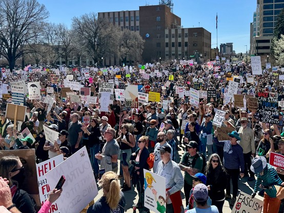 A crowd of protesters gathers in an urban setting, holding various signs with messages related to social and political issues. The scene is lively, with people of different ages and backgrounds participating in the demonstration under clear blue skies. Signs display slogans denouncing fascism.