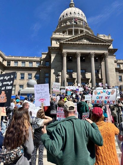 A large crowd gathers on the steps of the Idaho Capitol, holding various protest signs expressing political messages. The scene is lively with participants wearing casual clothing, and a clear blue sky is visible above. The architectural features of the building, including its dome.