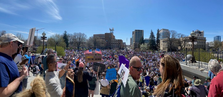 A large crowd gathers at an outdoor rally, holding various signs and banners. The scene features diverse participants, some visible in the foreground, with buildings and trees in the background under a clear blue sky. Signs convey messages of activism and political engagement.
