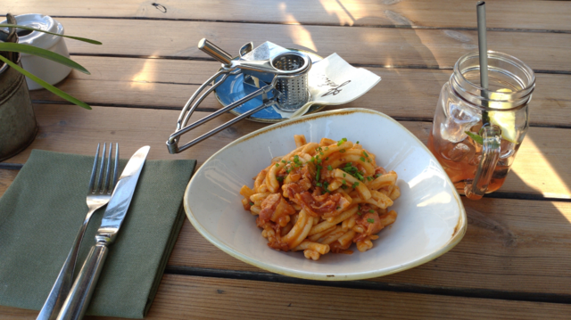 Pasta (hab vergessen, wie die Sorte hiess, jedenfalls kurze, längs seitlich gekringelte Pasta) mit Speck, Zwiebeln, Petersilie und angeblich Chili. Dahinter eine metallerne Käsereibe und rechts ein hausgemachter Eistee.