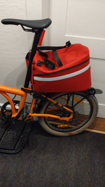 Das Heck eines orangefarbenen Brompton Electric G-Line mit einer roten, hinten runden Tasche mit weissem Reflektionsstreifen auf dem Gepäckträger, an der Sattelstange etwas zusammengedrückt.