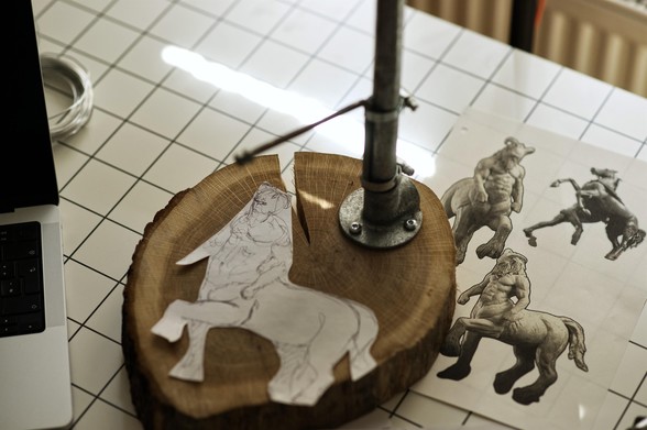 Photograph of a sculpting armature stand for clay sculpting, with printed out reference images next to it, and a sketch of the planned sculpture laying on top of it.