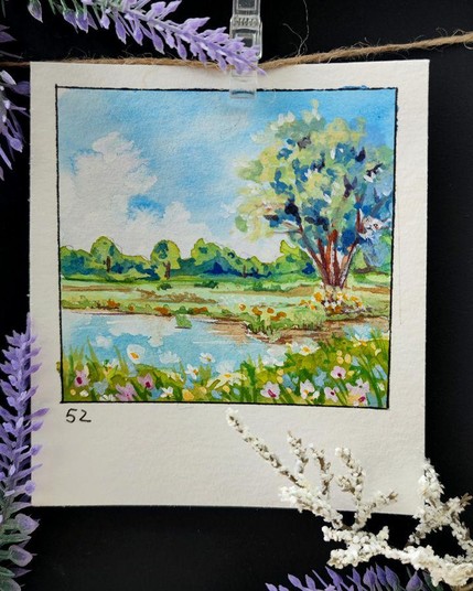 Quadratisches Aquarell, mit einer schwarzen Linie umrahmt, auf einem rechteckigen, hochformatigen Papier. Eine ruhige Flusslandschaft mit einem großen, buschigen Baum auf der rechten Seite, dessen Zweige sich in sanften Grün- und Blautönen gen Himmel strecken. Die Spiegelung des Baumes flimmert im ruhigen Wasser darunter. Im Vordergrund blühen verschiedene Wildblumen in Gelb, Weiß und Rosa entlang des Ufers und verleihen der Szene Lebendigkeit. Am Horizont erstrecken sich in sattem Grün entfernte Bäume und Büsche unter einem strahlend blauen Himmel mit fluffigen weißen Wolken. Die Zahl 52 ist in der unteren linken Ecke geschrieben.