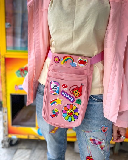 A person wearing a light-colored top and a pink jacket is shown from the waist up, featuring a pink utility-style fanny pack adorned with various colorful patches, including a smiling flower, rainbows, and playful phrases. The individual is also wearing distressed jeans.