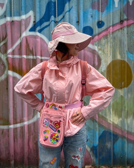 A person wearing a pink jacket, pink hat, and denim jeans stands against a colorful graffiti backdrop. The outfit features a waist pouch adorned with various colorful patches.