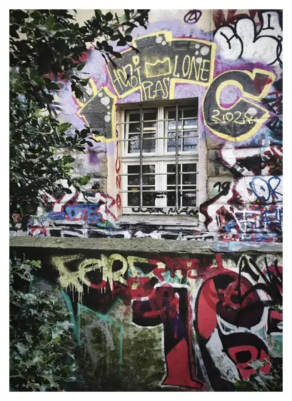 Blick über eine mit bunten Graffitti bemalte Mauer auf eine Hauswand, in der sich bildmittig ein großes viergeteiltes Fenster befindet. Das Fenster ist von außen mit jeweils 2 senkrechten und 2 waagerechten Eisenstäben vergittert. Der Bereich von bald mehr als einem Meter um das Fenster herum ist mit großen bunten Graffiti bemalt. Am linken Bildrand ragen grün belaubte Zweige eines Baumes ins Bild.