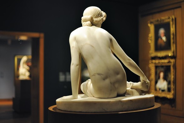a sculpture in marble of a young woman with her hair tied up with flowers in it, she is holding her foot, supporting her body weight with her left hand, she is seen from behind, in a museum , old paintings on the walls around her