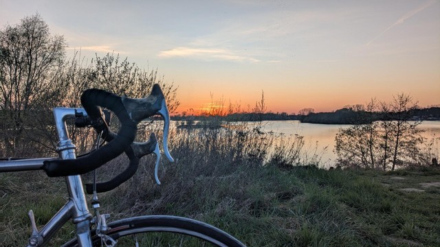 Sonnenuntergang an einem See, erhöhte Perspektive über der Uferböschung, im Vordergrund angeschnitten ein  Rennrad mit gebogenem Lenker