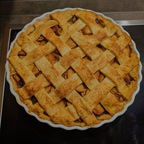 Goldbrauner Apfelkuchen mit Gitterkruste in einer weißen Keramikform, karamellisierte Apfelstücke sichtbar darunter.