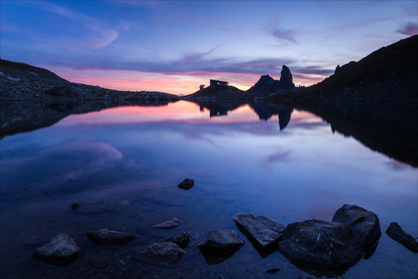 Photo d'un paysage de montagne au coucher du soleil.
Au premier plan, des cailloux baignent dans les eaux peu profondes d'un bord de lac. De l'autre côté du lac, un refuge se détache sur le ciel rose-orangé, juste à côté d'un pic montagneux dressé comme une canine. Ils se reflètent dans le lac.
-
Photo of a mountain landscape at sunset.
In the foreground, pebbles bathe in the shallow waters of a lakeshore. On the other side of the lake, a hut stands out against the pink-orange sky, right next to a canine-like mountain peak. They are reflected in the lake.