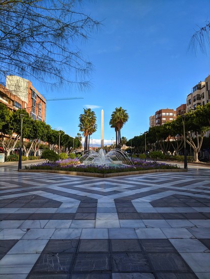 A wide, eye-level shot captures a vibrant urban scene under a clear, bright blue sky, likely in the Rambla de Almería. The foreground features a geometrically patterned pavement in shades of grey and white, leading towards a central fountain. The fountain is surrounded by a circular flower bed filled with purple flowers and lush green foliage, with water spraying upwards, catching the sunlight.
Behind the fountain, a tall, slender, light-coloured monument stands prominently. Flanking the fountain are two mature palm trees, their fronds reaching towards the sky. The middle ground is a pedestrian walkway lined with evenly spaced trees with green leaves, creating a verdant avenue. Street lamps with a classic design are placed along this walkway.
On either side of the walkway, multi-storey buildings in warm, earthy tones of orange and brown rise against the clear sky. Some buildings have balconies and multiple windows, indicating residential or commercial use.
