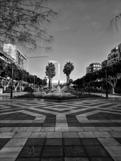A black and white, eye-level photograph captures a public square, likely the Rambla de Almería, under a cloudy sky. The foreground features a geometrically patterned pavement composed of light and dark tiles arranged in a central, star-like design that extends outwards. This paved area leads towards a central fountain, which is actively spraying water upwards and outwards, creating a dynamic visual element.
Surrounding the fountain is a circular area with dark foliage and some lighter-toned plants. Behind the fountain stands a tall, slender monument, appearing as a light vertical form against the darker backdrop. Flanking the fountain are two prominent palm trees with their characteristic fronds.
The middle ground consists of a pedestrian walkway lined with trees that have dense, dark foliage, creating a shaded avenue. Period-style street lamps are positioned along this walkway, their ornate designs visible in silhouette.