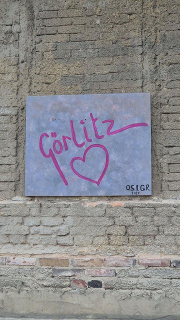 An einer rauen Backsteinwand hängt eine graue Metallplatte. Darauf steht in handgeschriebener pinker Schrift: „Görlitz“ mit einem großen Herzsymbol darunter. In der unteren Ecke ist „O.S.I.G.R. 2024“ zu lesen – vermutlich die Signatur der Künstler:innengruppe.