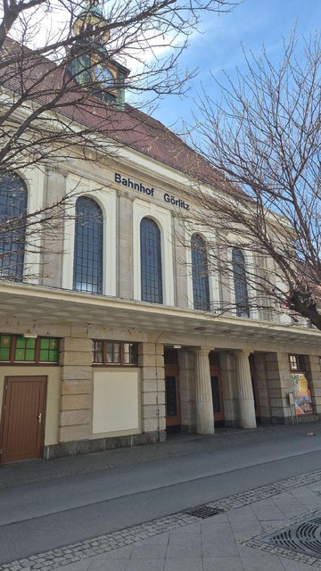 Das Bild zeigt die Vorderansicht des Bahnhofsgebäudes in Görlitz mit klassizistischer Architektur, hohen Rundbogenfenstern und dem Schriftzug „Bahnhof Görlitz“. Ein Uhrturm ragt über das rote Dach hinaus, im Vordergrund sieht man kahle Baumäste.
