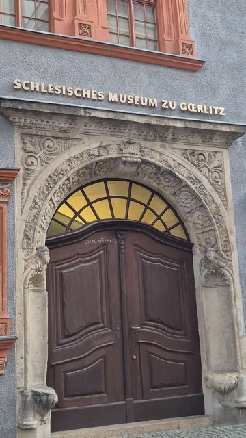 Das Eingangsportal des Schlesischen Museums in Görlitz zeigt ein großes dunkles Doppeltor mit barocker Steinrahmung. Oberhalb der Tür steht in goldener Schrift „Schlesisches Museum zu Görlitz“. Der Türbogen ist reich mit floralen und figürlichen Motiven verziert. Das Baujahr 1617 ist mittig im Bogen eingelassen.