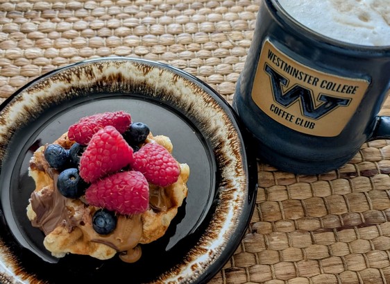 A Belgian waffle sits on a brown plate with raspberries, blueberries, Nutella, and speculoos on top. A blue coffee cup that says Westminster college coffee club with a big w sits beside it with milk froth on top. They sit on a wicker placemat.