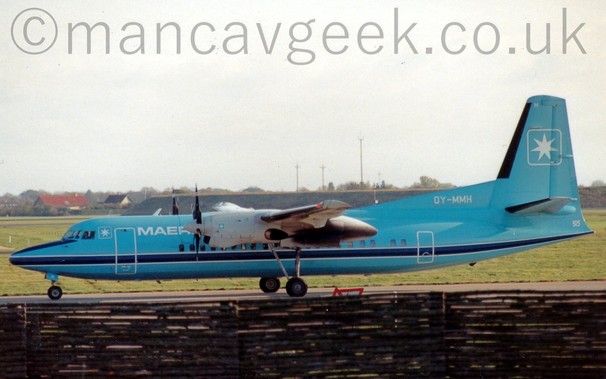 Side view of a light blue, high-winged, twin propellor-engined airliner taxiing from right to left.
The plane is mostly light blue, with a darker blue stripe outlined in white running along the body just below the passenger cabin windows.
There is the white outline of a square, with the corners rounded off, and containing a white, 7 pointed star, on the upper forward fuselage between the cockpit windows and the forward door, with white "Maersk Air" titles just aft of the door, although only the first 4 letters are visible, hidden behind the white engine pods poking out from the wings.
A larger version of the same square and star logo appears on the tail, with a smaller version on the side of the engine pods, just behind the propellors at the front.
The registration "OY-MMH" is on a fillet at the front of the tail, in white.
The main undercarriage is attached to open bays in the rear of the engine pods, on large retractable legs.
A brown wicker fence fills the foreground, the plane almost seeming to be taxiing along the top of it, with green grass in the background leading up to a grass covered berm in the background on the right, and red and brown roofs of houses on the left, under grey skies.
