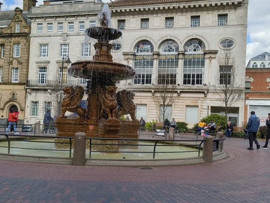 A victorian gothic fountain with three layers. The bottom layer is of four winged lions - three of them are visible. The fountain is in a greenish water circular pool with railings. There are white stone buildings behind. Some people are milling about in the photo.