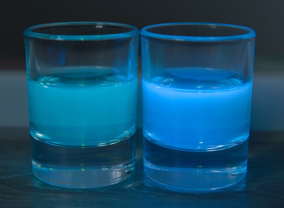 Zwei Schnapsgläser halb gefüllt im Tageslicht und mit UV-Licht beleuchtet. Links ist grünliche Floureszenz und rechts blaue zu sehen.