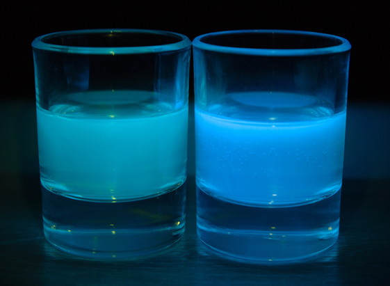 Zwei Schnapsgläser halb gefüllt im abgedunkelten Raum und mit UV-Licht beleuchtet. Links ist grünliche Floureszenz und rechts blaue zu sehen.