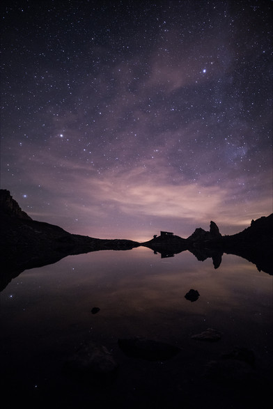 Photo d'un paysage de montagne nocturne au format portrait.
Un ciel très étoilé se reflète dans un lac de montagne. Le ciel a des teintes un peu violacées, quelques fins nuages apportent une ambiance fantomatique dans les étoiles.
Au tiers inférieur de l'image, un horizon composé d'un refuge et d'une pointe montagneuse en forme de canine ferme le lac dans un reflet impeccable.
-
Portrait photo of a mountain landscape at night.
A starry sky is reflected in a mountain lake. The sky has a slightly purplish tint, and a few thin clouds add a ghostly atmosphere to the stars.
In the lower third of the image, a horizon composed of a refuge and a canine-shaped mountain peak closes the lake in a flawless reflection.