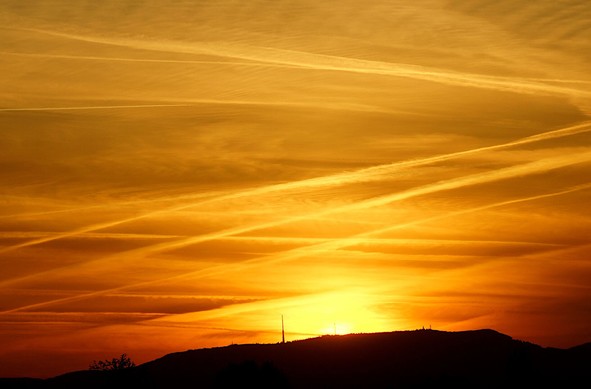 The silhouette of a mountain with some towers. The sun just rises behind the summit. The sky is orange, red, gray with clouds and crossing condensation strips.